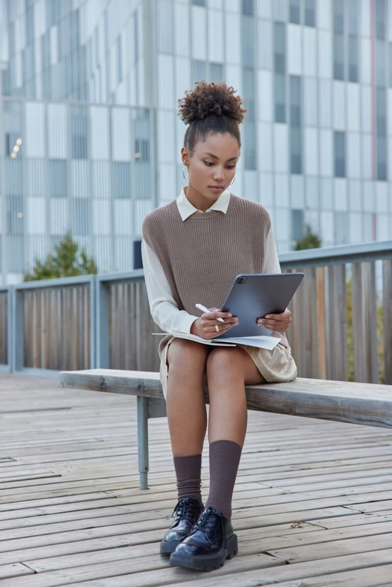 full-length-shot-curly-female-student-studies-outdoors-watches-webinar-via-digital-tablet-makes-notes-writes-down-necessary-information-sits-bench-against-modern-city-building-background (1)